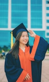 A young woman is wearing a navy blue and orange graduation gown and cap with a yellow tassel. She has long hair and is smiling broadly while adjusting her cap. The background features a modern building with large blue-tinted windows.