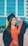 A young woman is wearing a navy blue and orange graduation gown and cap with a yellow tassel. She has long hair and is smiling broadly while adjusting her cap. The background features a modern building with large blue-tinted windows.