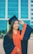 A young woman is wearing a navy blue and orange graduation gown and cap with a yellow tassel. She has long hair and is smiling broadly while adjusting her cap. The background features a modern building with large blue-tinted windows.