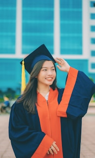 A young woman is wearing a navy blue and orange graduation gown and cap with a yellow tassel. She has long hair and is smiling broadly while adjusting her cap. The background features a modern building with large blue-tinted windows.