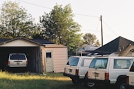A suburban scene with an old garage housing one SUV and three similar white SUVs parked outside. The garage and the vehicles have an aged appearance, with utility poles and trees in the background. Houses are visible, suggesting a residential neighborhood.