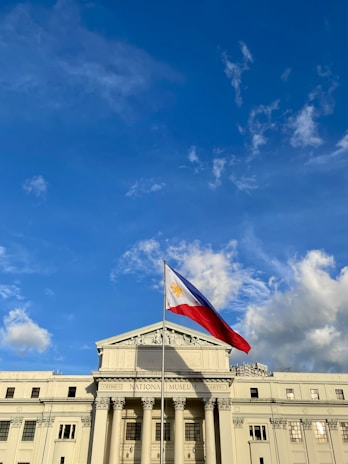 A grand neoclassical building with columns and a triangular pediment, prominently displaying the Philippine flag waving in the breeze against a clear blue sky with scattered clouds.