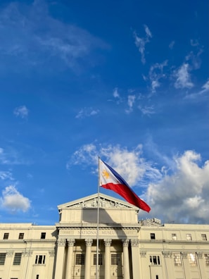 A grand neoclassical building with columns and a triangular pediment, prominently displaying the Philippine flag waving in the breeze against a clear blue sky with scattered clouds.