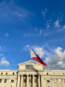 A grand neoclassical building with columns and a triangular pediment, prominently displaying the Philippine flag waving in the breeze against a clear blue sky with scattered clouds.
