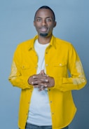 A person stands against a plain blue background, wearing a bright yellow button-up shirt over a white t-shirt. Their hands are clasped together at chest level, and they have a relaxed and approachable demeanor.