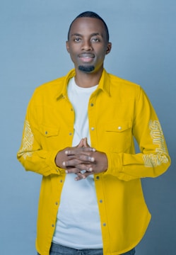 A person stands against a plain blue background, wearing a bright yellow button-up shirt over a white t-shirt. Their hands are clasped together at chest level, and they have a relaxed and approachable demeanor.