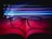 An open book is positioned with a pair of black-rimmed glasses resting on its pages. Colorful, neon light streaks of blue, purple, and pink pass through the air above, creating a dynamic play of light and shadow on the pages.