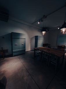 A dimly lit room with industrial-style lighting and minimal furnishings. There are two large, teal-colored cabinets against the wall. A wooden table with metal chairs is set in the center of the room, creating a functional yet sparse appearance.
