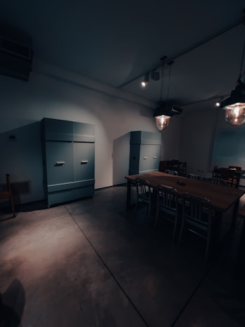 A dimly lit room with industrial-style lighting and minimal furnishings. There are two large, teal-colored cabinets against the wall. A wooden table with metal chairs is set in the center of the room, creating a functional yet sparse appearance.