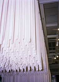 A large installation composed of various white vertical panels or strips hanging from the ceiling in an indoor, industrial-style space. The panels are evenly spaced and create a rhythmic, geometric pattern.