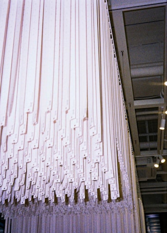 A large installation composed of various white vertical panels or strips hanging from the ceiling in an indoor, industrial-style space. The panels are evenly spaced and create a rhythmic, geometric pattern.