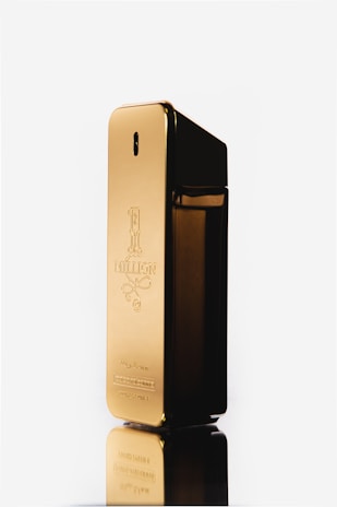 A gold rectangular perfume bottle with a reflective surface and embossed text on the front. The bottle stands upright against a white background, showcasing its sleek and luxurious design.