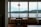 A cozy café interior featuring a small table with two wooden chairs and a modern lamp above. Large windows provide a scenic view of a calm body of water and distant green hills under a cloudy sky.