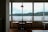 A cozy café interior featuring a small table with two wooden chairs and a modern lamp above. Large windows provide a scenic view of a calm body of water and distant green hills under a cloudy sky.