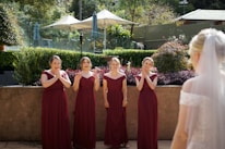 A group of four bridesmaids in matching burgundy dresses stand together outdoors, displaying expressions of joy and surprise. They are looking towards a bride in a white dress with a veil, who is partially visible from the back. The setting is lush and green with hedges, plants, and a patio area, suggesting a garden or outdoor venue.
