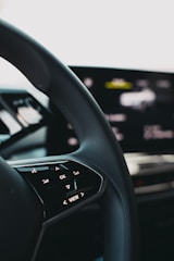 A close-up view of a car steering wheel, featuring control buttons including 'OK' and directional arrows. The background shows a blurred digital display indicating technological elements.
