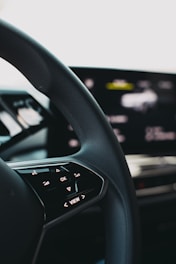 A close-up view of a car steering wheel, featuring control buttons including 'OK' and directional arrows. The background shows a blurred digital display indicating technological elements.