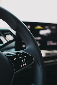 A close-up view of a car steering wheel, featuring control buttons including 'OK' and directional arrows. The background shows a blurred digital display indicating technological elements.