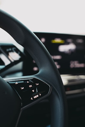 A close-up view of a car steering wheel, featuring control buttons including 'OK' and directional arrows. The background shows a blurred digital display indicating technological elements.