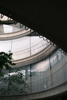 A modern architectural structure with curved glass windows and vertical metal railings reflecting light. A tree is visible in the foreground, adding natural contrast to the sleek design.