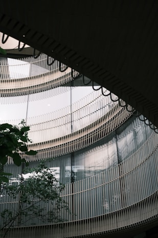 A modern architectural structure with curved glass windows and vertical metal railings reflecting light. A tree is visible in the foreground, adding natural contrast to the sleek design.
