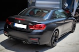 A sleek, black BMW M4 is parked outdoors in a well-lit area. The car features a glossy finish and distinct red taillights, with dual exhaust pipes visible at the rear. The vehicle is positioned in a way that highlights its sporty and elegant design. In the background, there is a motorcycle partially visible.