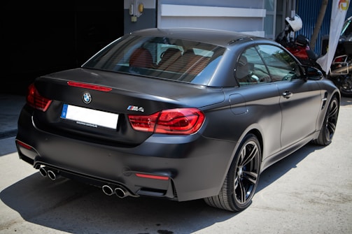 A sleek, black BMW M4 is parked outdoors in a well-lit area. The car features a glossy finish and distinct red taillights, with dual exhaust pipes visible at the rear. The vehicle is positioned in a way that highlights its sporty and elegant design. In the background, there is a motorcycle partially visible.