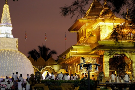 A large, ornate golden temple is brightly illuminated against the evening sky, with intricate architectural details and designs. Near the temple is a stupa adorned with numerous small lights, creating a delicate glow. People, dressed in white garments, gather around these structures, suggesting a religious or cultural event. Palms and other decorative elements enhance the scene.