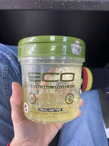 A hand holding a clear plastic container of Eco Style Professional Styling Gel with Olive Oil. The container is slightly transparent, revealing the gel inside. It prominently features the product details, including 'Alcohol-Free' and 'For All Hair Types.' The cap is green, and text on the container notes benefits like repairs, conditions, and moisturizes. In the background, there is a blurred view of denim fabric and a sock with red and yellow stripes.