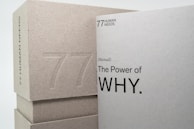 Two books or boxes are stacked, with the visible text '77 Human Needs' and '(Manual) The Power of WHY.' embossed on the surfaces. The materials appear to be made of a textured, grayish paper or cardboard.
