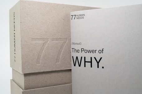 Two books or boxes are stacked, with the visible text '77 Human Needs' and '(Manual) The Power of WHY.' embossed on the surfaces. The materials appear to be made of a textured, grayish paper or cardboard.