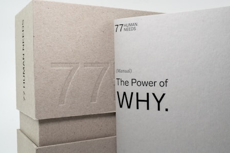 Two books or boxes are stacked, with the visible text '77 Human Needs' and '(Manual) The Power of WHY.' embossed on the surfaces. The materials appear to be made of a textured, grayish paper or cardboard.