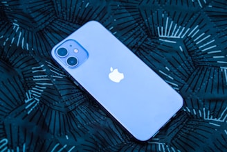 A smartphone with a prominent logo is lying on a dark patterned surface. The phone has a smooth, sleek design with dual rear cameras.