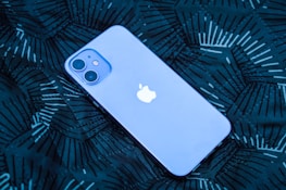 A smartphone with a prominent logo is lying on a dark patterned surface. The phone has a smooth, sleek design with dual rear cameras.