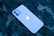 A smartphone with a prominent logo is lying on a dark patterned surface. The phone has a smooth, sleek design with dual rear cameras.