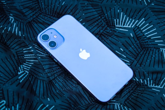 A smartphone with a prominent logo is lying on a dark patterned surface. The phone has a smooth, sleek design with dual rear cameras.