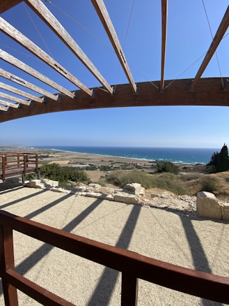 A wooden pergola offers partial shade with beams casting long shadows on a gravel patio. Beyond, a vast landscape stretches out, featuring fields and rolling hills that meet the blue horizon of the sea. Vegetation and rocky patches are scattered across the land, and the sky is clear with no clouds.