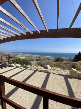 A wooden pergola offers partial shade with beams casting long shadows on a gravel patio. Beyond, a vast landscape stretches out, featuring fields and rolling hills that meet the blue horizon of the sea. Vegetation and rocky patches are scattered across the land, and the sky is clear with no clouds.