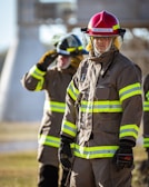 Firefighters wearing protective gear, including helmets and uniforms with reflective stripes, stand in an outdoor setting with a focused demeanor.