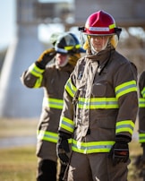Firefighters wearing protective gear, including helmets and uniforms with reflective stripes, stand in an outdoor setting with a focused demeanor.