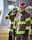 Firefighters wearing protective gear, including helmets and uniforms with reflective stripes, stand in an outdoor setting with a focused demeanor.