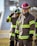 Firefighters wearing protective gear, including helmets and uniforms with reflective stripes, stand in an outdoor setting with a focused demeanor.