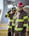 Firefighters wearing protective gear, including helmets and uniforms with reflective stripes, stand in an outdoor setting with a focused demeanor.