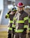Firefighters wearing protective gear, including helmets and uniforms with reflective stripes, stand in an outdoor setting with a focused demeanor.