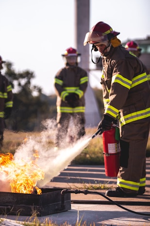 Firefighters in gear actively participate in a training exercise, using fire extinguishers to control and suppress a controlled fire. Protective helmets and reflective stripes on uniforms are visible, emphasizing safety protocols.