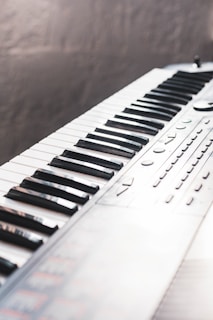 A close-up of a musical keyboard, showing an array of black and white keys with buttons and control knobs on the side. The keyboard is positioned diagonally in the frame, creating a dynamic perspective.