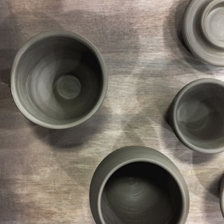 Several unfinished clay pots are placed on a wooden surface. The pots appear to be of varying sizes, showcasing a raw, earthy texture. The lighting casts subtle shadows, highlighting the smooth, rounded edges of the pottery.