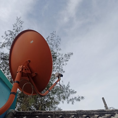 A large orange satellite dish mounted on a roof with trees in the background under a partly cloudy sky.