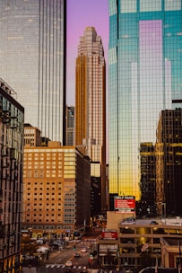 A cityscape featuring tall skyscrapers with reflective glass facades under a gradient sky transitioning from purple to orange. The view includes a busy urban street with a self-parking sign and various vehicles moving through the intersection.