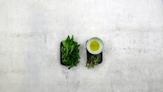 On a textured light gray surface, two dark rectangular trays contain fresh green herbs and leaves. To the right of the trays, a white bowl holds a yellow liquid, likely olive oil.
